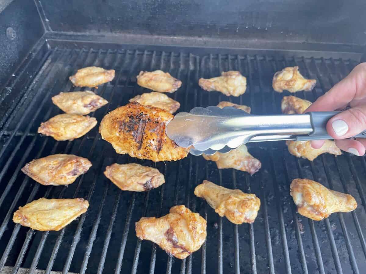 a pair of tongs holding up a partially cooked chicken wing with grill marks above a grill with an even layer of cooking chicken wings
