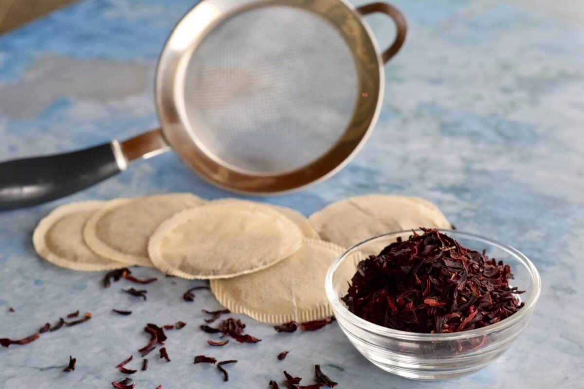 a small strainer for loose leaf tea on a blue board backdrop with 5 tea bags and a small bowl of loose leaf hibiscus tea