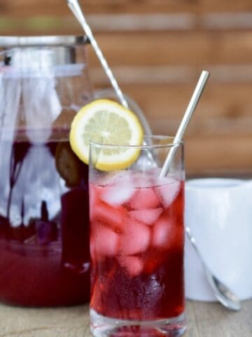 glass pitcher filled with berry hibiscus iced tea next to a glass with poured iced and a stirring spoon and a small white pitcher in front of a wood slat background