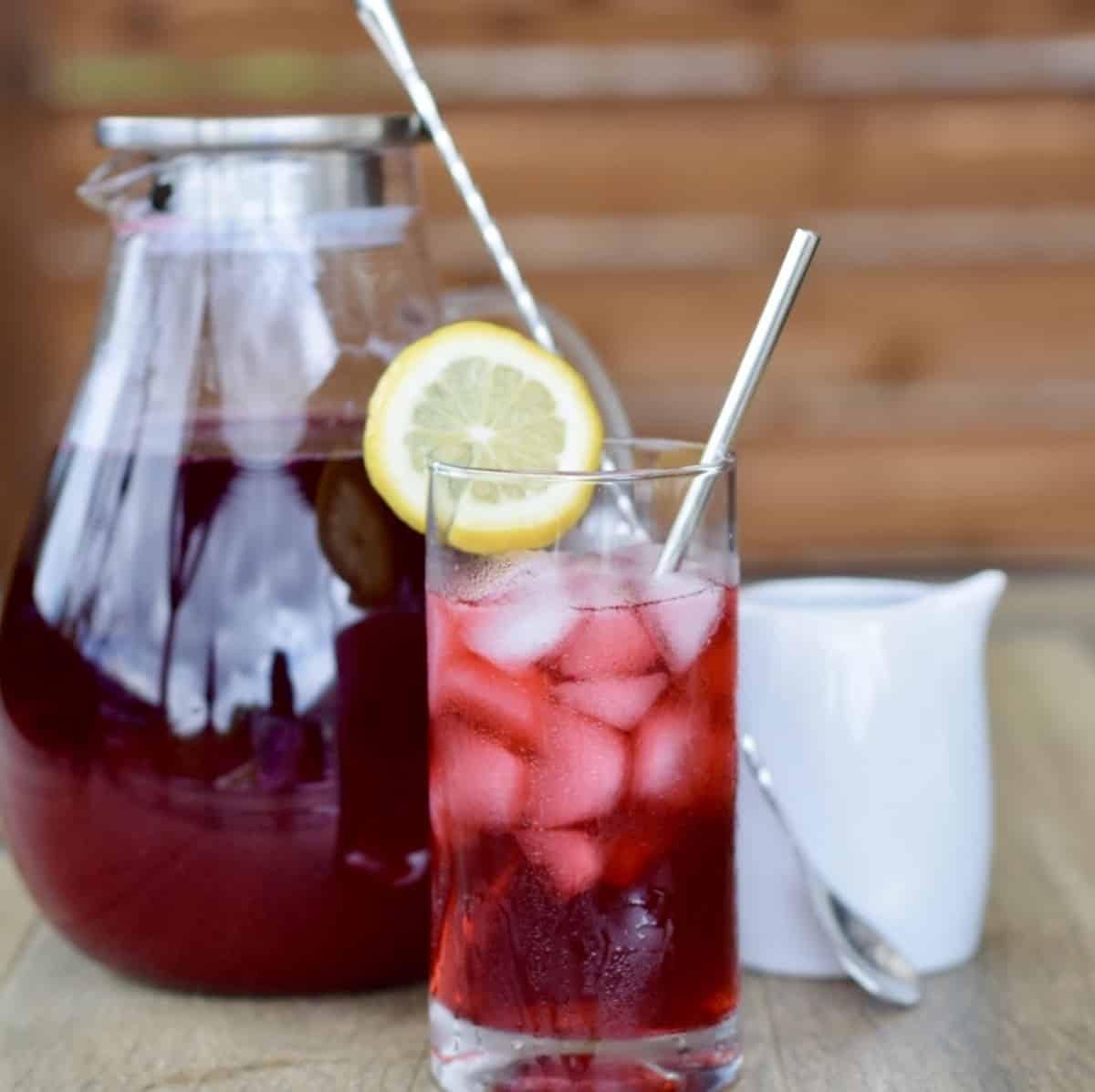 glass pitcher filled with berry hibiscus iced tea next to a glass with poured iced and a stirring spoon and a small white pitcher in front of a wood slat background
