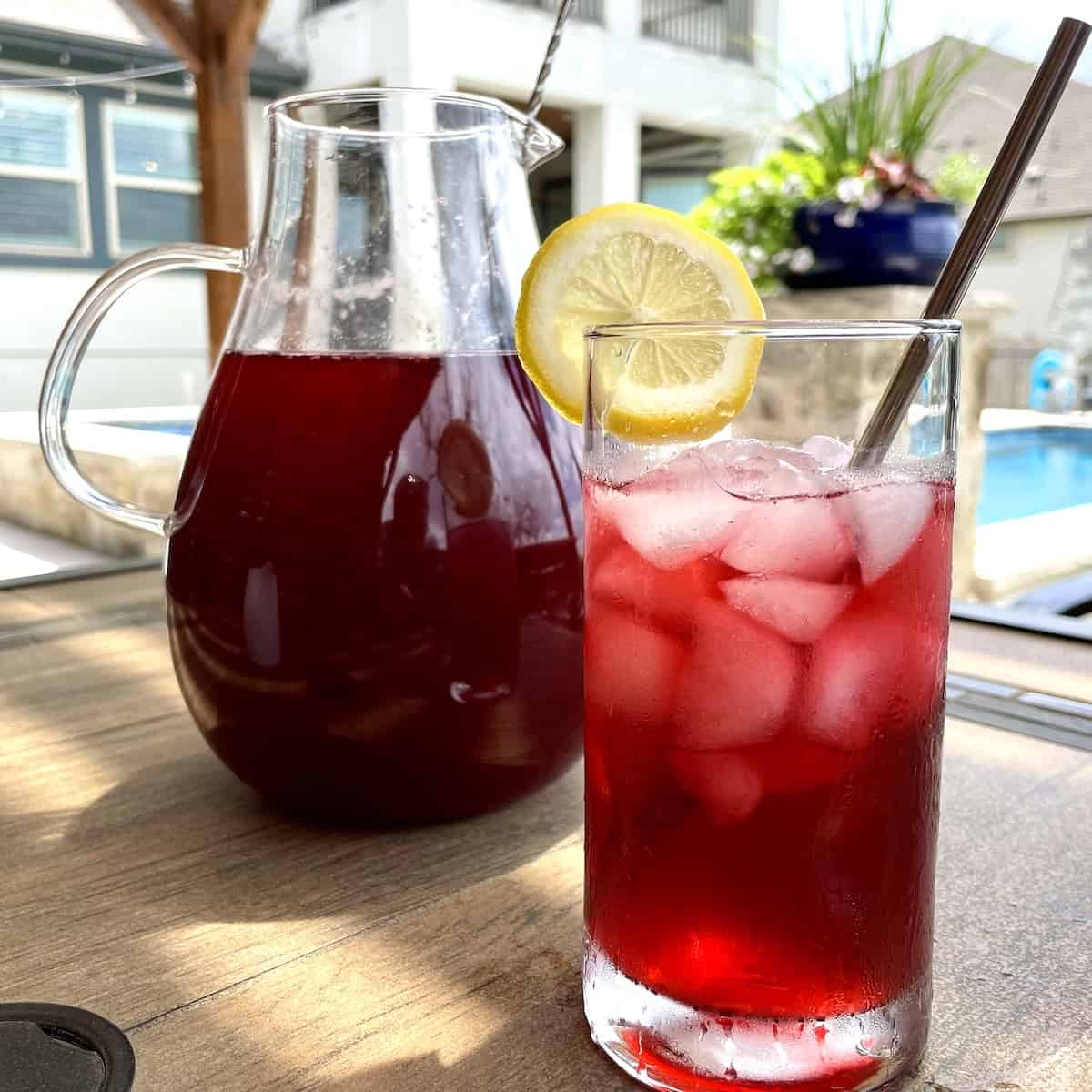 a glass pitcher filled with berry hibiscus iced tea on a brown slate table with a full drinking glass of iced tea with a straw and garnished with a lemon