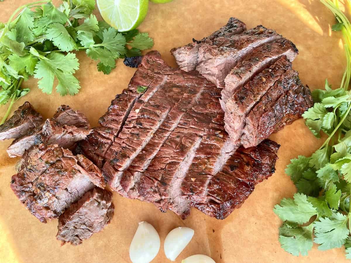 a brown cutting board with cooked traeger carne asada sliced with cilantro and garlic cloves for garnish