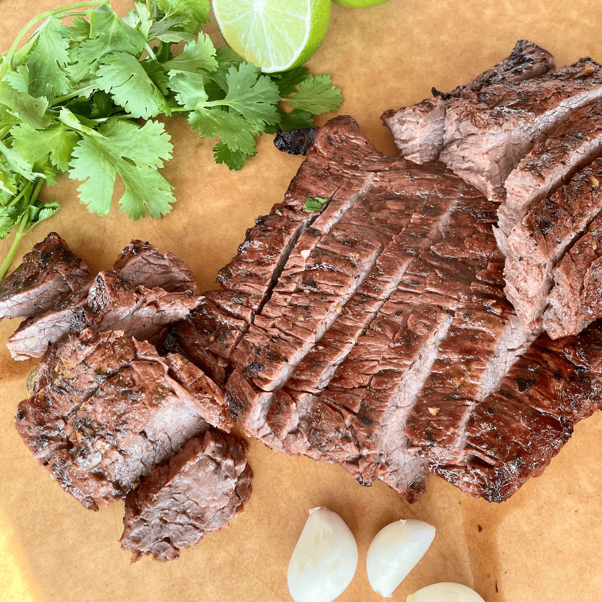 sliced cooked traeger carne asada on a brown cutting board with fresh cilantro cut lime and garlic cloves