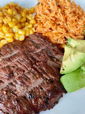 a white plate with sliced carne asada mexican rice diced avocado and paprika seasoned corn