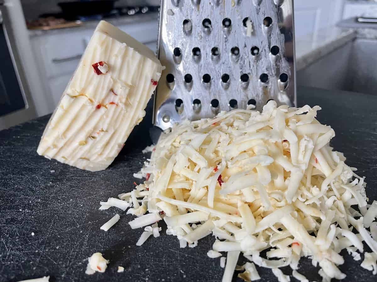 a large box cheese grater with a block of pepper jack cheese and large pile of shredded pepper jack cheese in front of it on a black cutting board