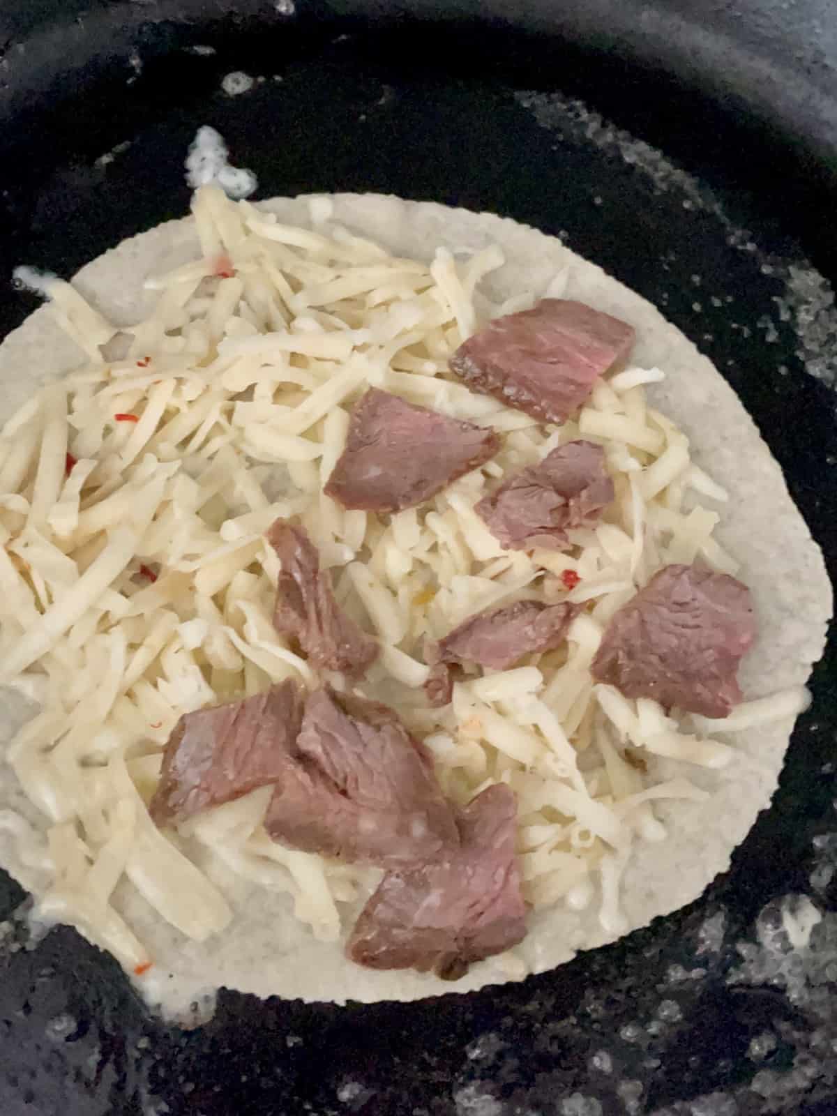 cast iron skillet with a carne asada quesadilla cooking open face showing the shredded cheese and carne asada