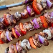 3 cooked chicken kabobs placed diagonally on a brown cutting board with metal tongs