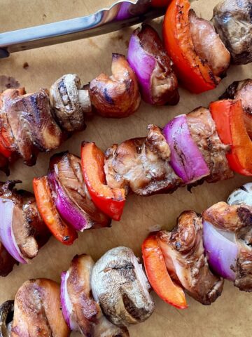 3 cooked chicken kabobs placed diagonally on a brown cutting board with metal tongs