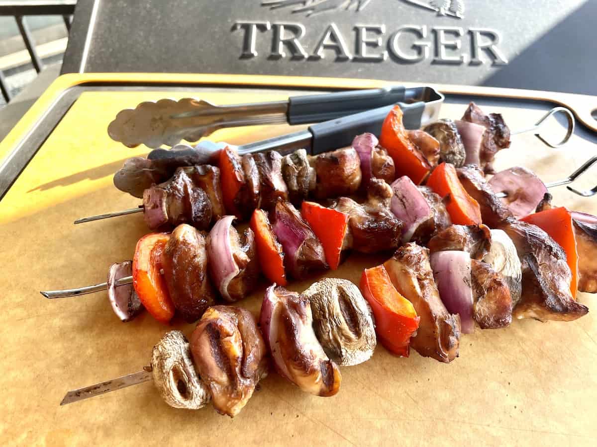 3 cooked chicken and vegetable kabobs on a large cutting board with tongs in front of a traeger grill