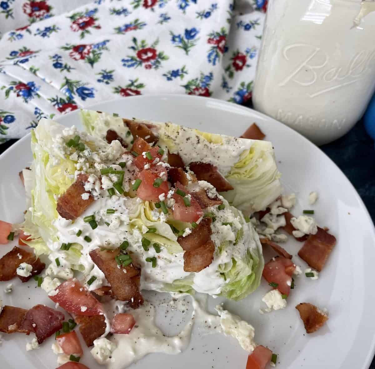 a wedge salad served on a white plate with a flower apron and jar of fresh blue cheese dressing in the background