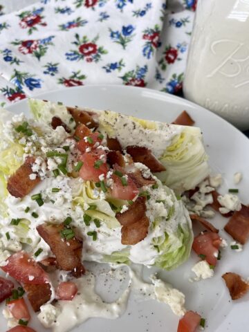 a wedge salad served on a white plate with a flower apron and jar of fresh blue cheese dressing in the background