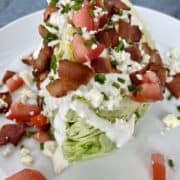 a white plate on a blue background holding a wedge of iceberg lettuce topped with homemade blue cheese dressing crumbled bacon diced tomatoes chopped chives and cracked black pepper