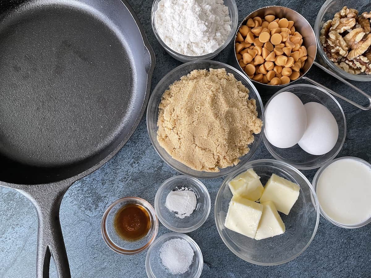 a 9 inch cast iron skillet next to ingredients for butterscotch blondies including flour butter eggs vanilla baking powder salt vanilla walnuts and cream on a blue background 