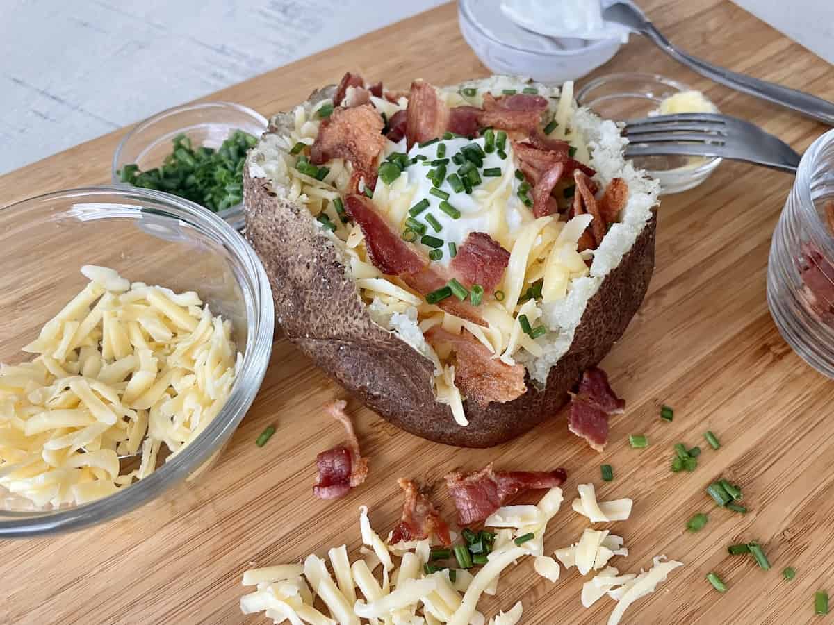 a traeger baked potato on a brown cutting board topped with butter sour cream crispy bacon shredded gouda cheese and diced chives
