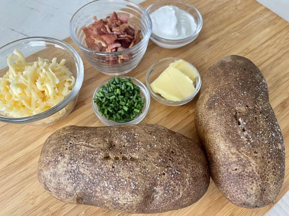 a brown cutting board with two salted traeger baked potatoes next to small dishes of shredded gouda cheese crispy bacon diced chives butter and sour cream