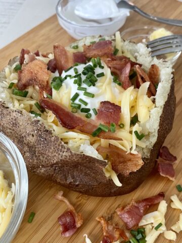 a baked potato topped with butter sour cream shredded smoked gouda cheese crispy bacon and diced chives on a wood cutting board and extra toppings sprinkled around the baked potato