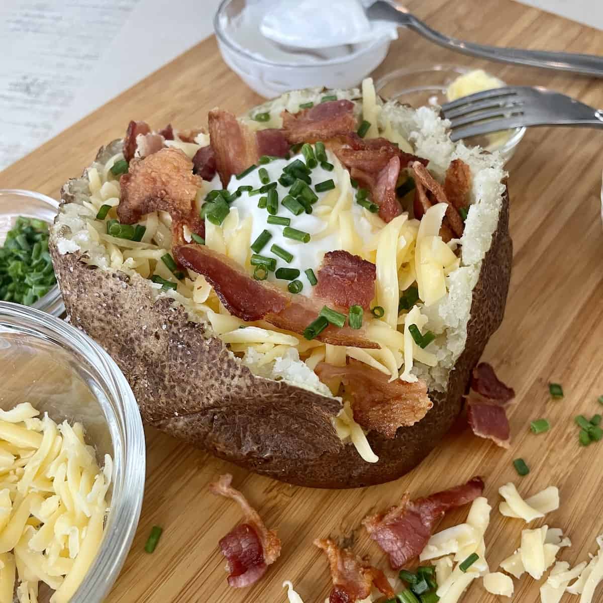 a baked potato topped with butter sour cream shredded smoked gouda cheese crispy bacon and diced chives on a wood cutting board and extra toppings sprinkled around the baked potato