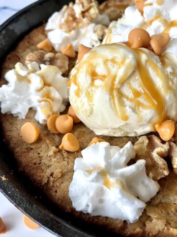 a baked butterscotch and walnut blondie in a cast iron skillet with scoops of vanilla ice cream topped with whipped cream caramel sauce and more butterscotch chips