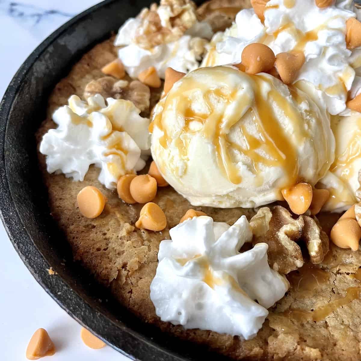 a baked butterscotch and walnut blondie in a cast iron skillet with scoops of vanilla ice cream topped with whipped cream caramel sauce and more butterscotch chips
