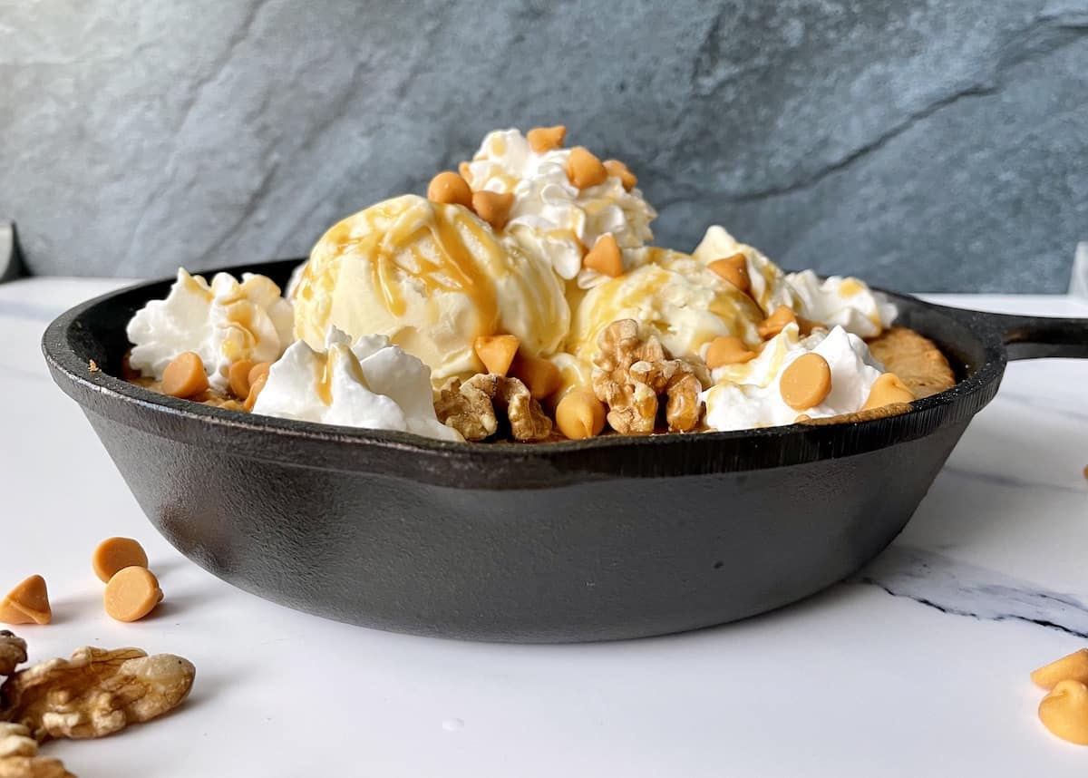 a cast iron pan with a blondie topped with vanilla ice cream whipped cream caramel sauce and butterscotch chips placed on a marble countertop with a blue backdrop and extra butterscotch chips scattered around the board