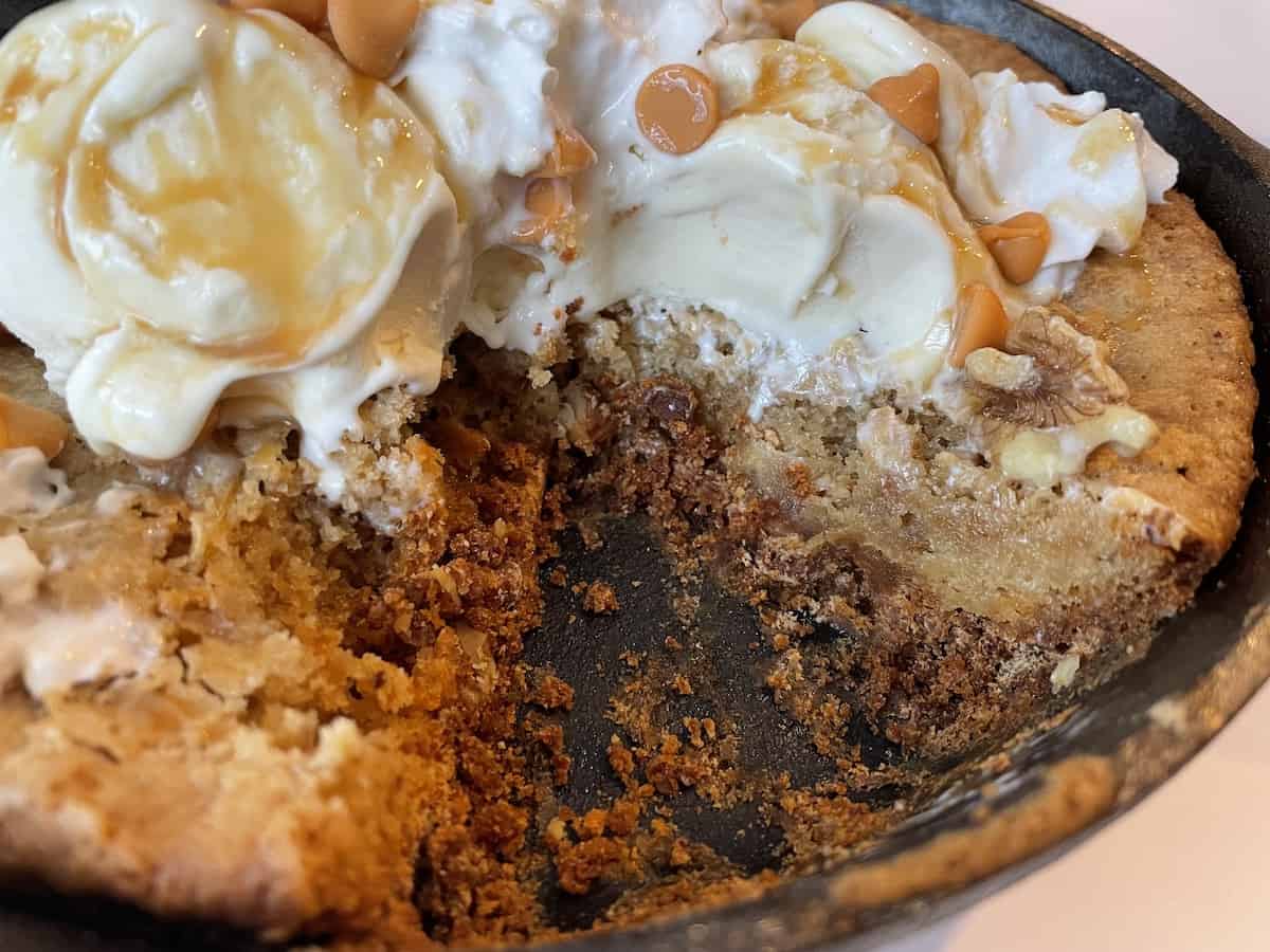 a butterscotch blondie in a cast iron pan with vanilla ice cream whipped cream and caramel sauce missing a portion which has been cut out