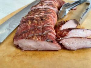 sliced traeger pork tenderloin on a light wood cutting board next to a meat knife and tongs