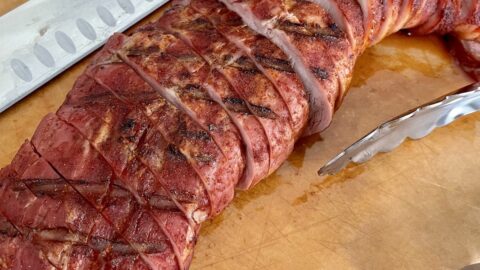 sliced pork tenderloin with a red smoky char served on a cutting board next to tongs and a meat slicing knife