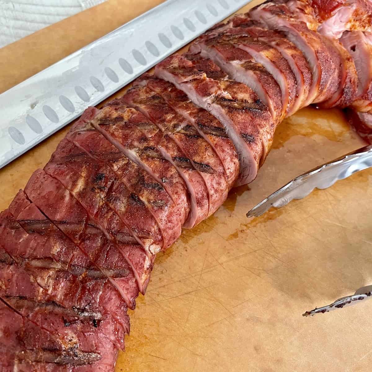 sliced pork tenderloin with a red smoky char served on a cutting board next to tongs and a meat slicing knife