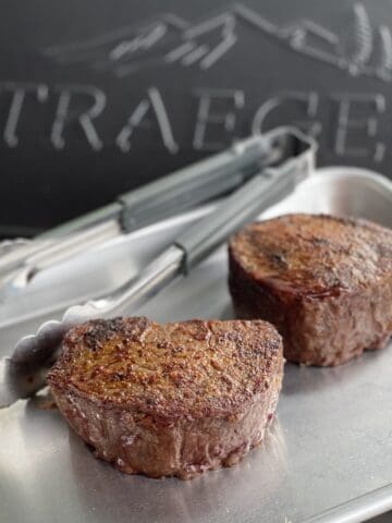 two perfectly cooked traeger filet mignon on a quarter sheet pan with tongs in front of a closed traeger grill