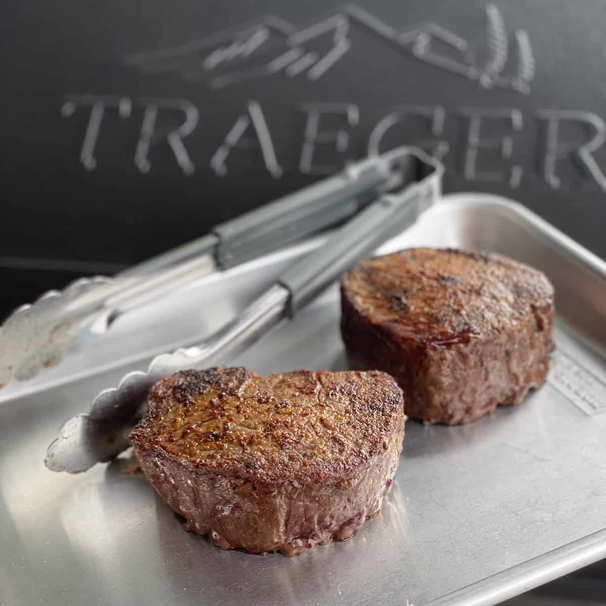 two perfectly cooked traeger filet mignon on a quarter sheet pan with tongs in front of a closed traeger grill