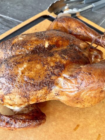 a dark brown smoked whole chicken resting on a light brown cutting board with silver tongs and a meat thermometer in the background