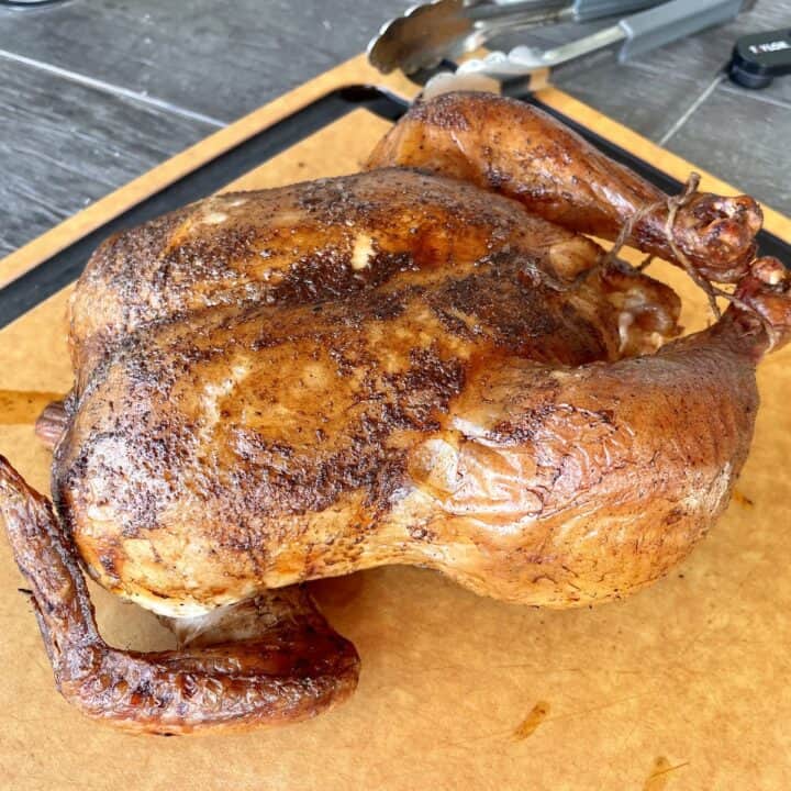 a dark brown smoked whole chicken resting on a light brown cutting board with silver tongs and a meat thermometer in the background