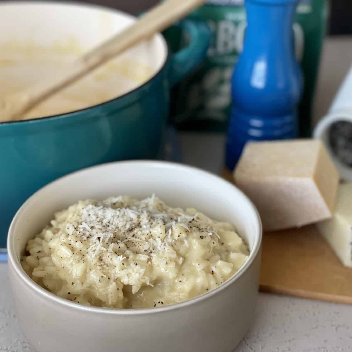 cacio e pepe risotto served in a tan bowl topped with fresh cracked black pepper and extra pecorino roam cheese with blocks of cheese a teal pot and blue pepper grinder in the background