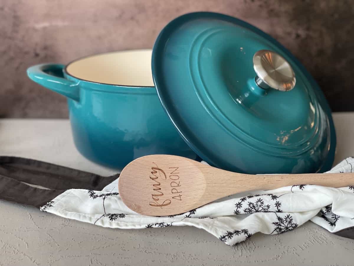 a teal dutch oven with the lid tilted on its side with a spoon that reads fancy apron on the end and a brown and white apron in front of a dark brown background 