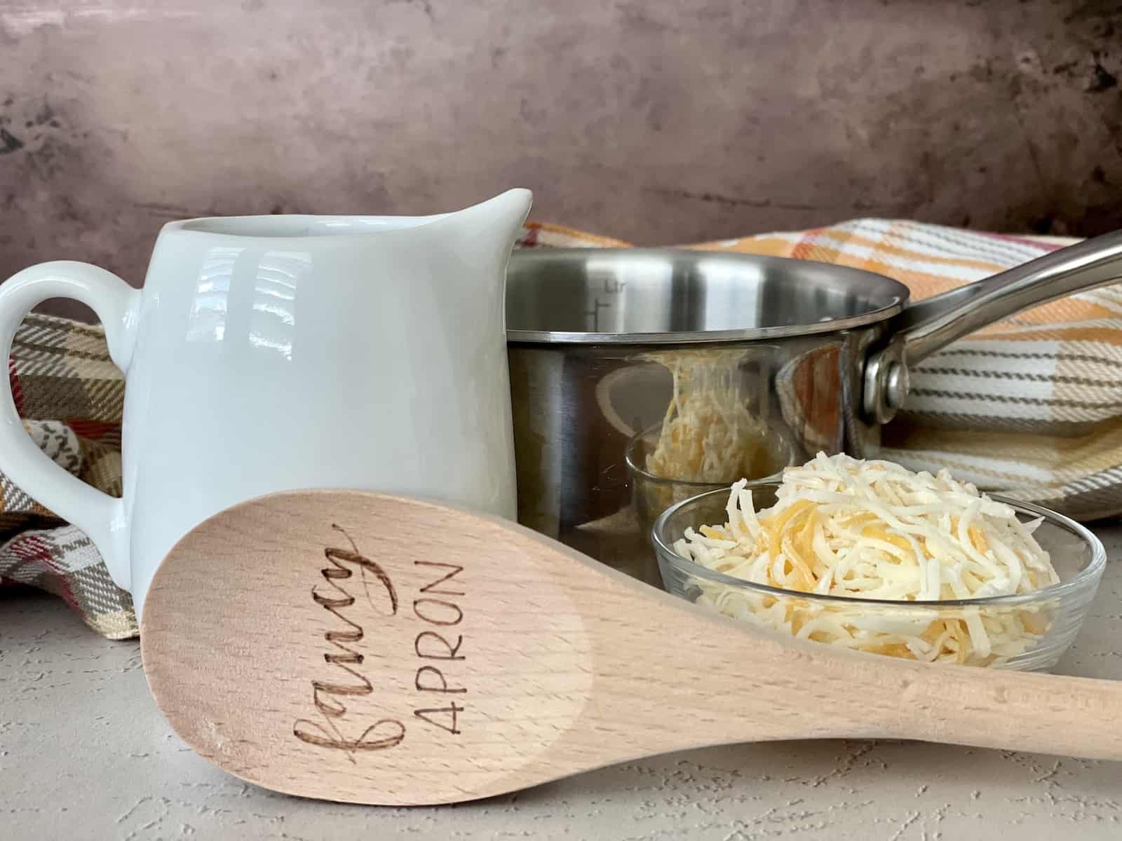 flourless cheese sauce ingredients including heavy cream in a white pitcher bowl of shredded cheese a small sauce pan a brown apron and a wood spoon that has fancy apron etched on it