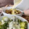 flourless cheese sauce pouring over a bowl of broccoli with a bowl of potatoes and a brown apron in the background