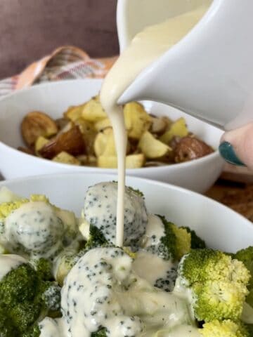 flourless cheese sauce pouring over a bowl of broccoli with a bowl of potatoes and a brown apron in the background
