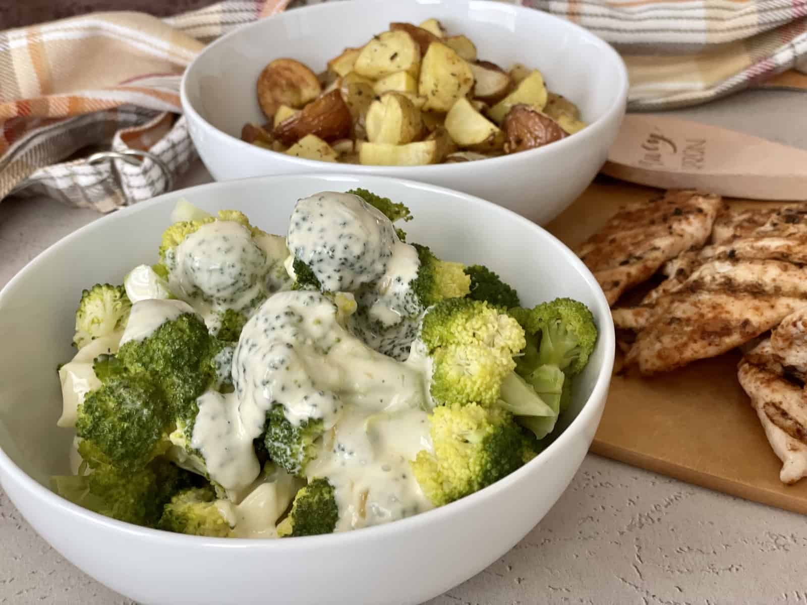 a bowl of bright green broccoli with flourless cheese sauce poured over served next to a bowl of roasted potatoes and grilled chicken on a wood cutting board