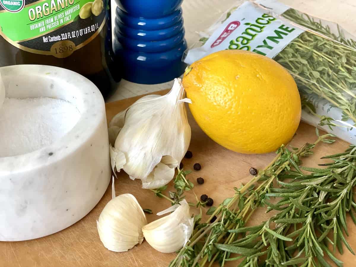 lemon and herb marinade ingredients including cloves of garlic a whole lemon marble jar of salt fresh rosemary and thyme a blue pepper grinder and bottle of olive oil