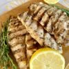 4 grilled chicken breast cutlets that have been marinated in lemon and herb marinade on a brow cutting board with extra thyme and rosemary and lemons cut in half for garnish