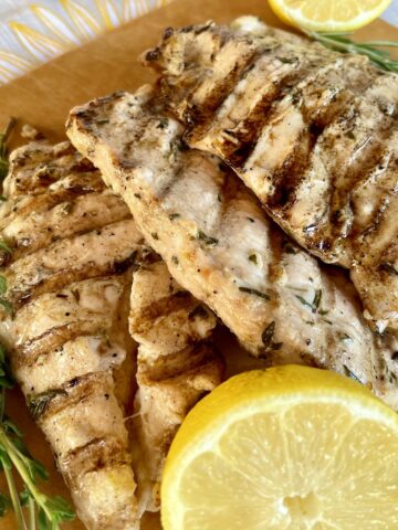 4 grilled chicken breast cutlets that have been marinated in lemon and herb marinade on a brow cutting board with extra thyme and rosemary and lemons cut in half for garnish