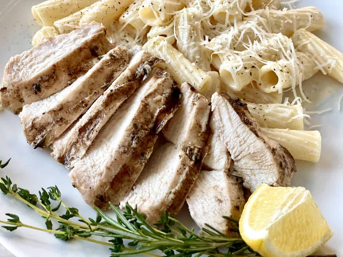 sliced marinated chicken served on a white plate with rigatoni and fresh herbs for garnish and a lemon wedge