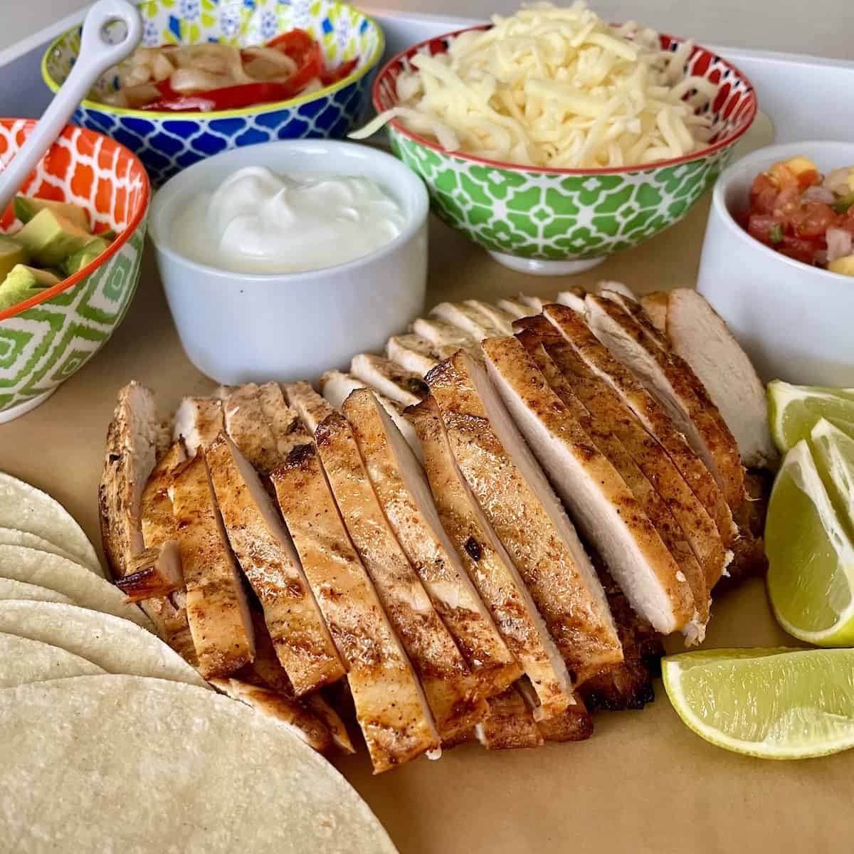 a large platter serving sliced tequila lime chicken with sour cream diced avocado fajita bell peppers and onions shredded cheese and mango salsa in colorful bowls