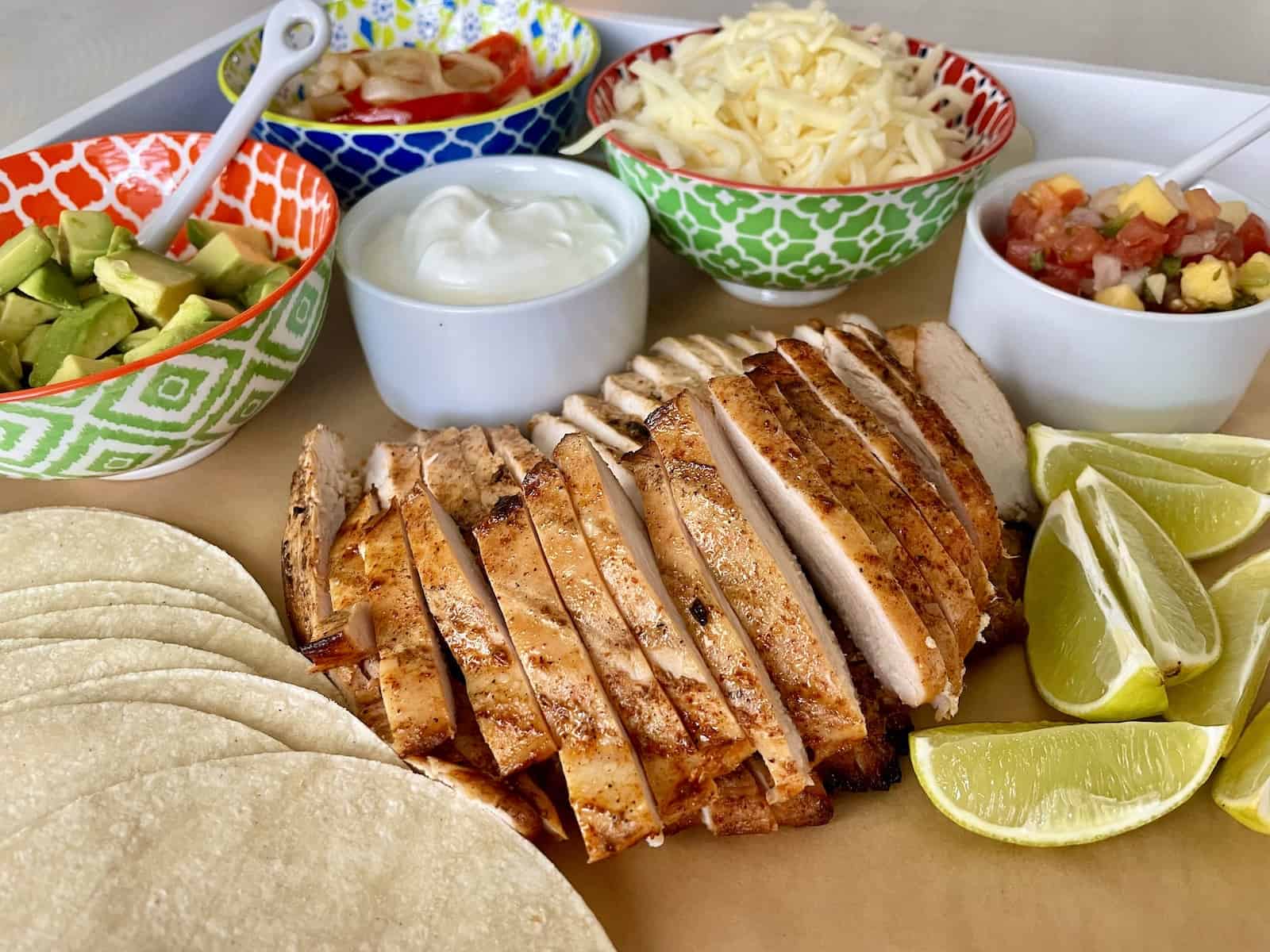 a large platter serving sliced tequila lime chicken with sour cream diced avocado fajita bell peppers and onions shredded cheese and mango salsa in colorful bowls