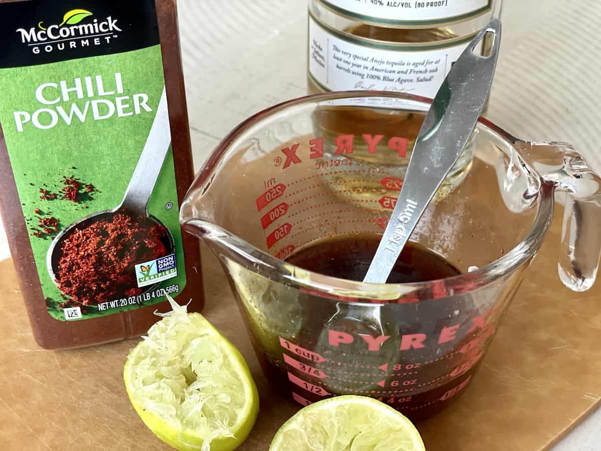 a measuring cup of prepared tequila lime chicken marinade on a cutting board with cut limes a bottle of tequila and jar of chili powder