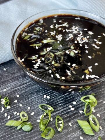 a glass bowl of soyaki sauce topped with green onions and sesame seeds on a grey wood background with a green kitchen towel