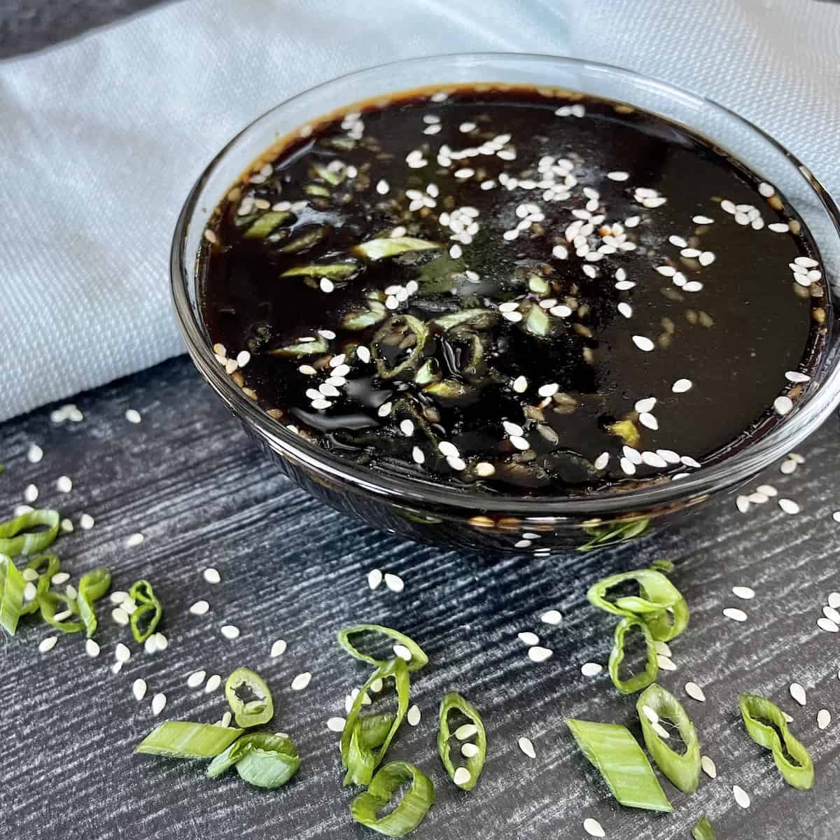 a glass bowl of soyaki sauce topped with green onions and sesame seeds on a grey wood background with a green kitchen towel