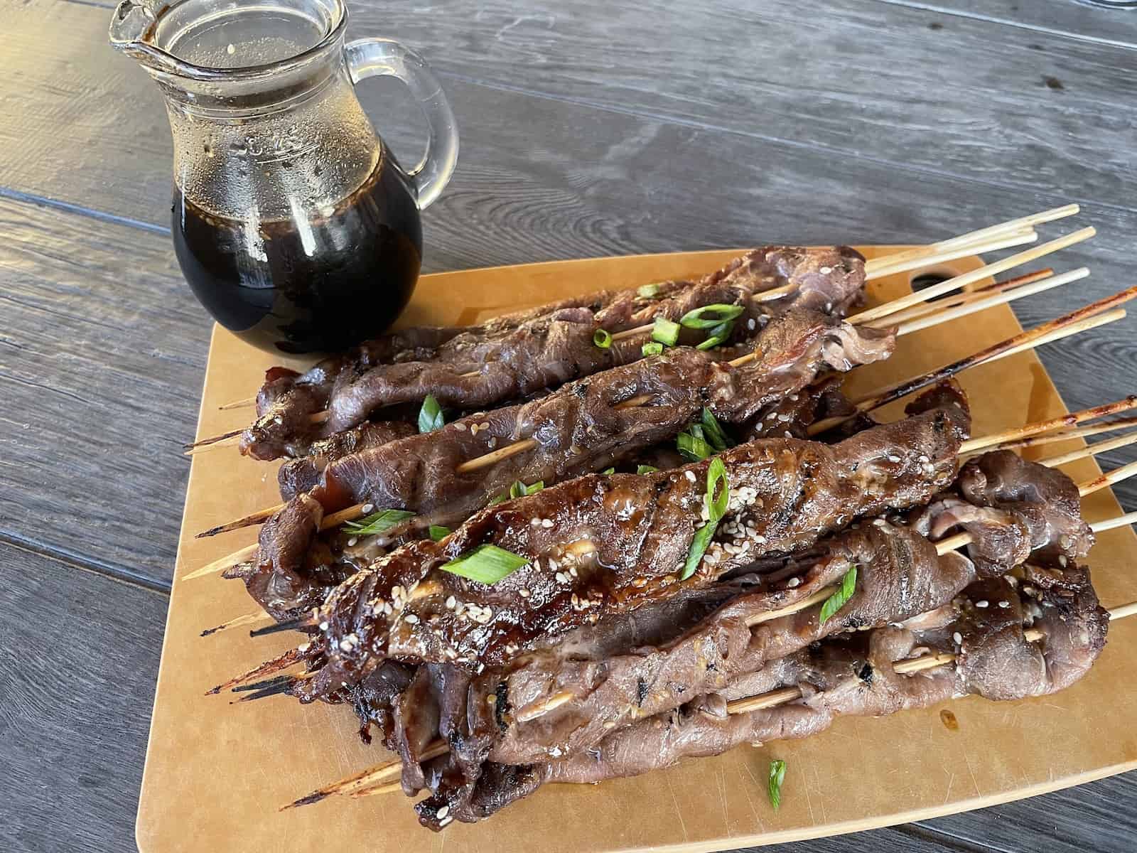 teriyaki steak skewers off the grill on a cutting board with a tiny pitcher of sauce on the side