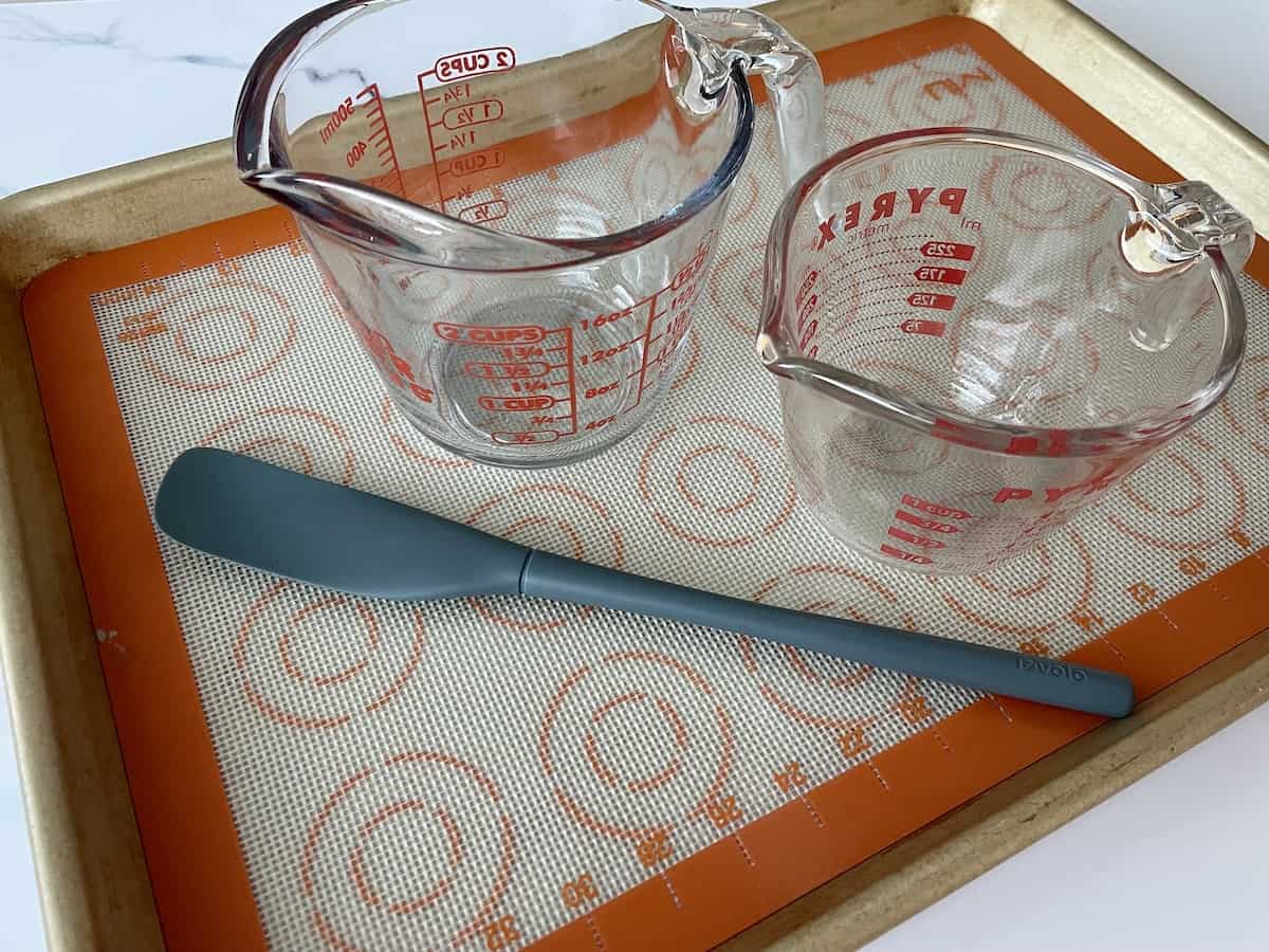 a gold baking sheet lined with a silpat two glass measuring bowls and a grey rubber spatula