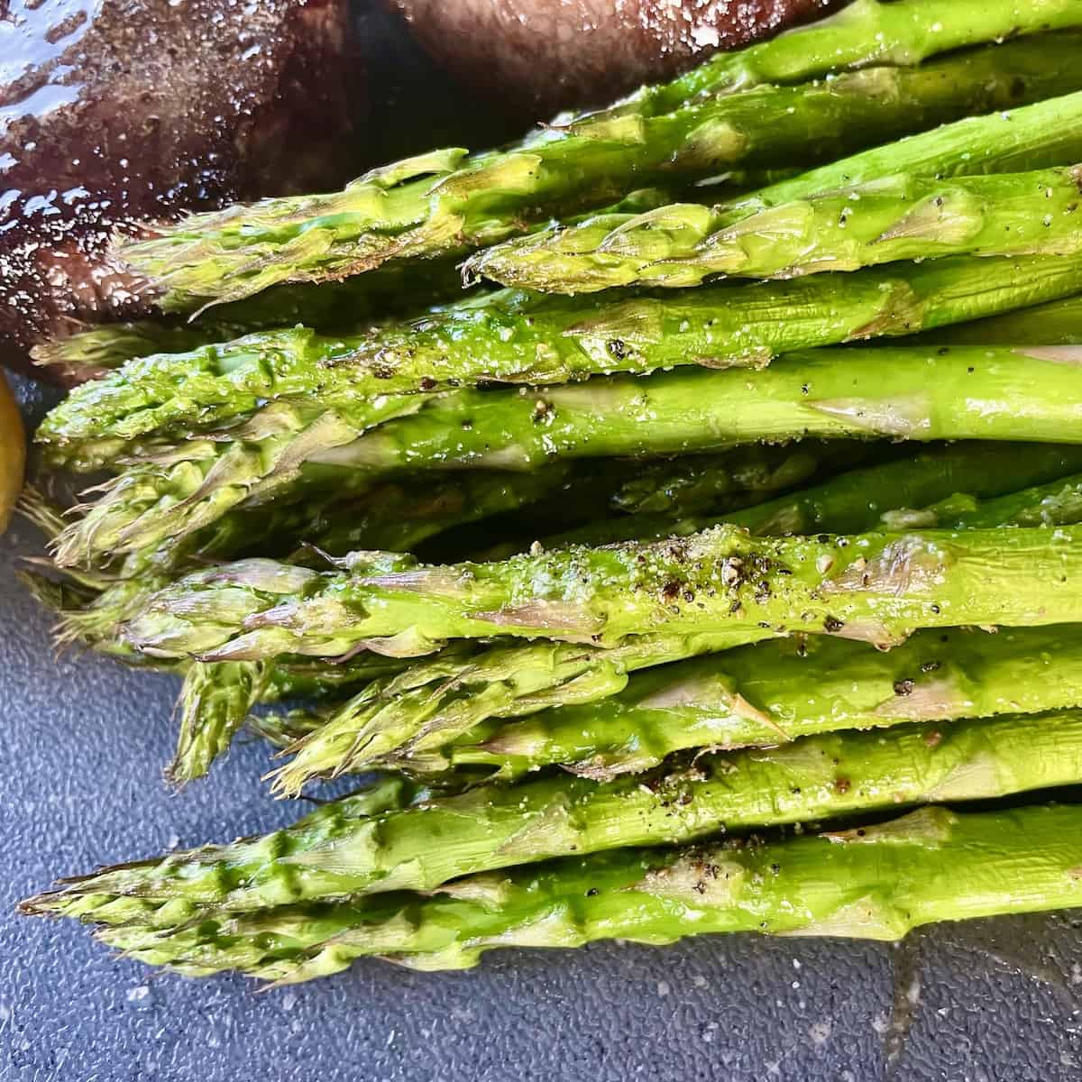a large pile of bright green grilled asparagus seasoned with salt and pepper on a black cutting board